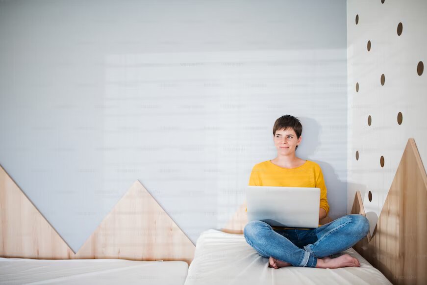 The image shows a person seated on the bed with a laptop on her legs