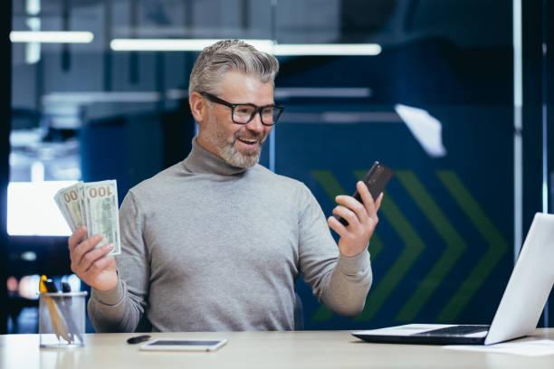 The image shows a person holding a mobile phone in one of his hands and money in the other hand